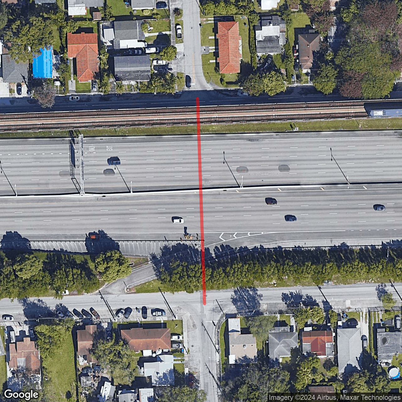 Aerial satellite image of a highway and rail corridor dividing two residential neighborhoods, with a red line marking the severed street connection